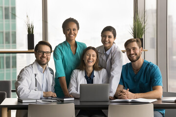 Diverse team of happy young doctors sitting and standing at laptop, looking at camera with toothy...
