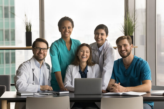 Diverse team of happy young doctors sitting and standing at laptop, looking at camera with toothy smiles, posing for group medic professional portrait. Medical practitioners promoting healthcare