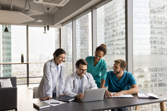 Young Multiethnic Business Team Talking To Colleagues On Online Video Conference Call, Sitting And Standing At Laptop, Looking At Screen Together, Laughing, Enjoying Medical Communication On Internet