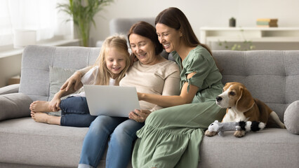 Three female generations of happy family and adorable dog resting on couch, using app, modern...
