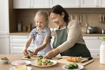 Positive senior grandma in kitchen apron teaching little granddaughter kid to cook, preparing...