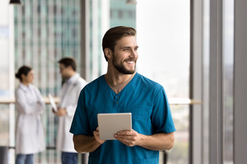 Happy handsome doctor man in blue uniform holding digital tablet, looking away, thinking, smiling, laughing, standing in clinic hall space, using work application, electronic online communication