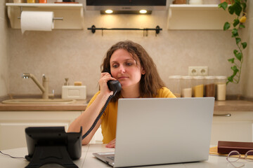 A woman with a laptop is talking on a landline phone at a table in a home kitchen. An adult female businesswoman works from home, a remote office - obrazy, fototapety, plakaty