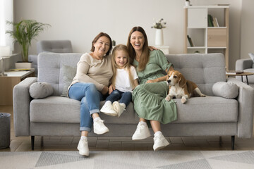 Happy girls and women of three family generations sitting at cute dog on comfortable couch close...