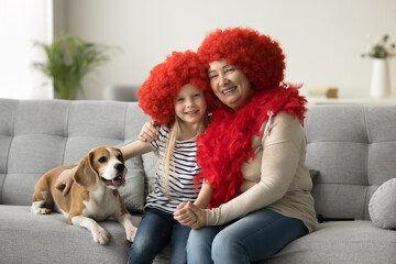 Happy little granddaughter and grandma in clown wigs stroking dog, sitting on couch, laughing,...