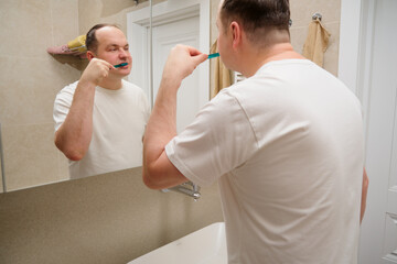 A man brushes his teeth with a toothbrush in the bathroom in front of the mirror. Overweight 40-year-old man (forty)