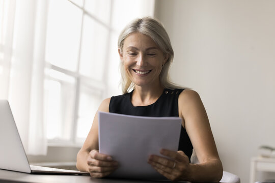 Attractive middle-aged woman sit at desk, hold paper letter, bank statement or notification, do paperwork, satisfied with financial report, get good news, feel happy, loan approval, investment income
