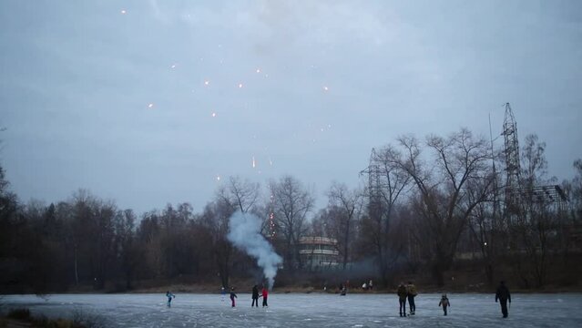 People on natural ice rink among trees launch fireworks at evening