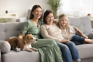 Happy beautiful mom, granny, little kid girl and dog resting on comfortable couch together, looking away, posing for family portrait, smiling, laughing, having fun, enjoying meeting at home