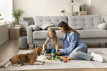 Positive caring mom and little kid playing with building blocks near dog on heating floor at home,...