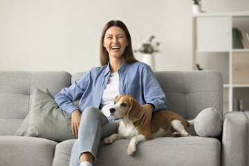 Happy joyful dog owner woman resting on comfortable stylish couch, stroking beagle at home, enjoying leisure time with cute beloved pet, looking at camera with toothy smile, laughing
