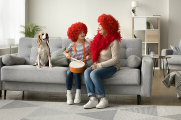 Cheerful little kid girl and grandma in red wigs sitting on couch at trick dog, playing toy drum,...