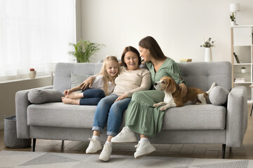 Wide family home portrait of three female generations with dog. Happy grandma, mother and little kid girl stroking cute beagle, sitting on sofa close together, talking, laughing