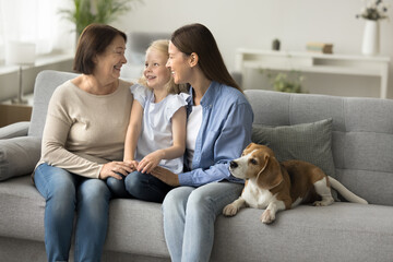 Happy mother, grandma and little child girl sitting on couch at adorable beagle dog, talking,...
