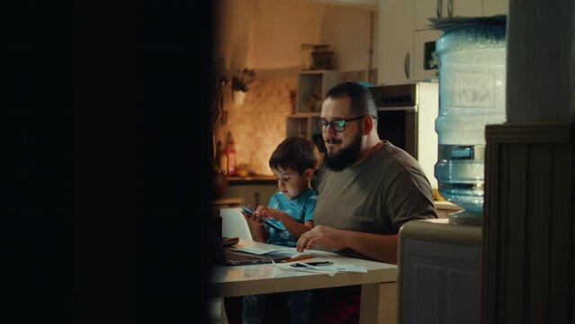 A father paying bills online using a laptop in the kitchen evening and holding his son on lap who playing games on a mobile phone
