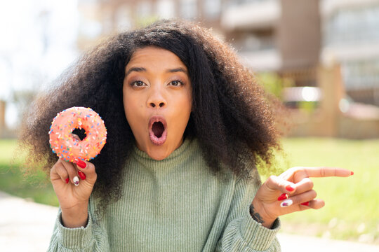 Young African American woman holding a donut at outdoors surprised and pointing side