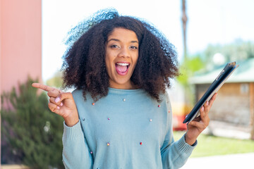 Young African American woman holding a tablet at outdoors surprised and pointing finger to the side