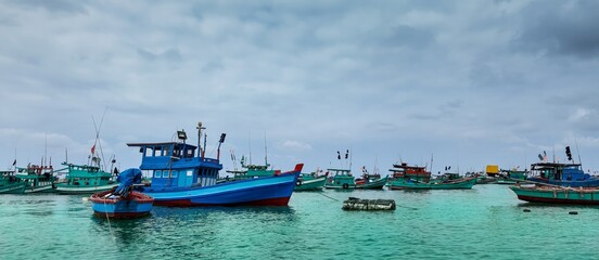 Fototapeta premium Colorful fishing boats anchored in a calm turquoise bay under an overcast sky, depicting a tranquil coastal fishing village scene