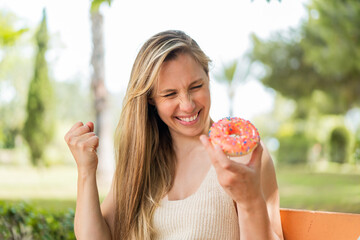Young blonde woman holding a donut at outdoors celebrating a victory