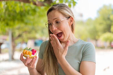 Young blonde woman holding a tartlet at outdoors with surprise and shocked facial expression - obrazy, fototapety, plakaty