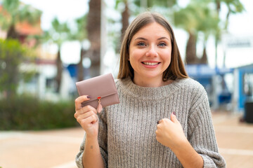 Young pretty blonde woman holding a wallet at outdoors with surprise facial expression