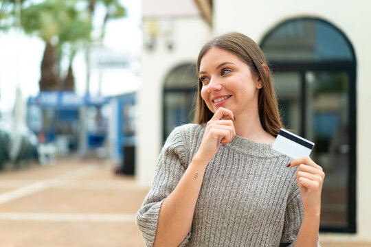 Young pretty blonde woman holding a credit card at outdoors thinking an idea and looking side