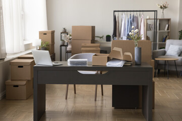 Cozy workplace with desk and stacked heap of cardboard boxes, hanger with clothes, no people. Moving day to new office, workspace of designer, selling fashion attire through internet, small business