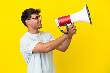 Young caucasian handsome man isolated on yellow background shouting through a megaphone to announce something - obrazy, fototapety, plakaty