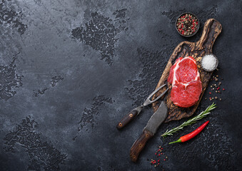 Raw rib eye steak fillet on wooden board with fork and knife,salt and pepper on dark kitchen table...