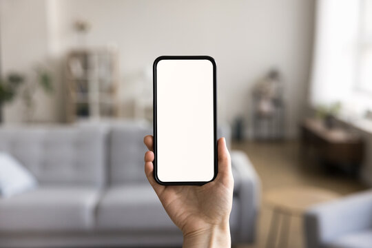 Close up shot of unknown female hand holding modern smartphone with white mock up blank screen, living room on background. Advertisement of new mobile application ad, smart home app, comfort living