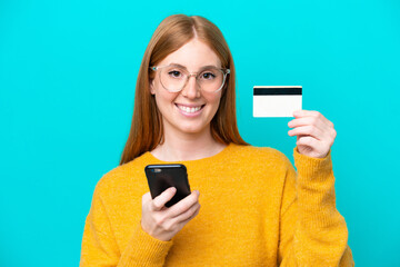 Young redhead woman isolated on blue background buying with the mobile with a credit card