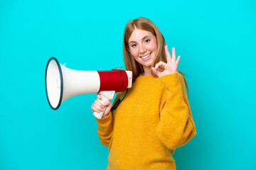 Young redhead woman isolated on blue background holding a megaphone and showing ok sign with fingers