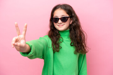 Little caucasian girl wearing sunglasses isolated on pink background smiling and showing victory sign