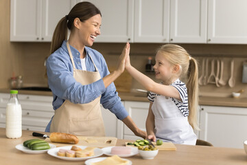 Cheerful proud mom teaching adorable little kid girl to cook, giving high five over table with...