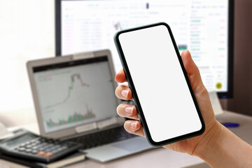 Smart phone with blank screen in hand in front of computers with financial charts on monitors.