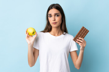 Young caucasian woman isolated on blue background having doubts while taking a chocolate tablet in...