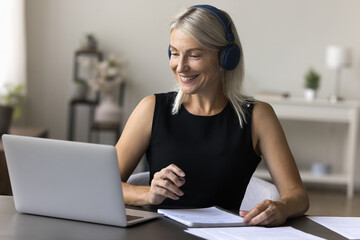 Happy pretty mature woman in headphones sit at desk, take notes, watch webinar on laptop, enjoy web...