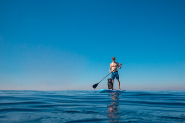 A man paddles on a Stand Up Paddle, SUP board with a black Brittany spaniel dog on the sea. Active...