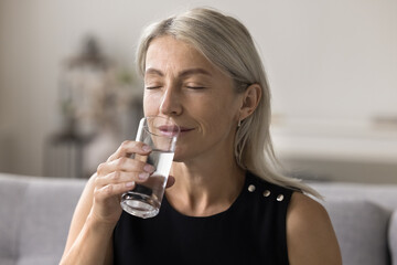 Close up shot of peaceful middle-aged woman holds glass, drinking clean, natural and filtered...