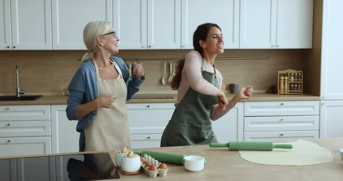 Cheerful excited blonde senior mom and adult daughter woman having fun in home kitchen, dancing to retro disco music, smiling, laughing, enjoying activity at table with roller, dough