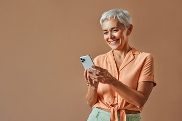 A beautiful stylish middle-aged woman stands on a beige background and holds a white smartphone...