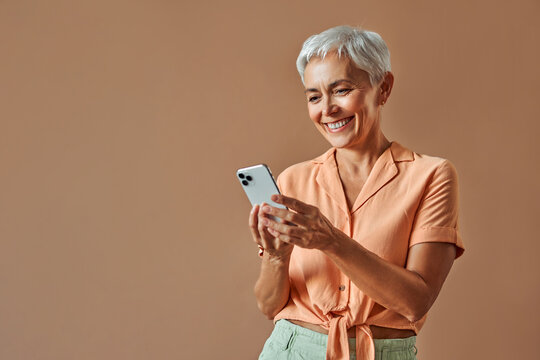 A beautiful stylish middle-aged woman stands on a beige background and holds a white smartphone with a smile. Advertising concert. Copy space. - Powered by Adobe