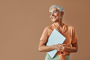 Confident beautiful mature gray haired stylish modern creative woman wearing glasses orange shirt and pastel green pants holding laptop and looking away to free space on beige background.