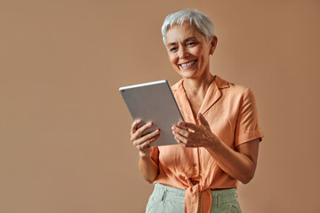 Smiling happy sincere confident mature beautiful gray haired stylish modern creative woman in pastel orange shirt and green pants holding digital tablet while standing on beige background.