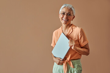 Mature beautiful confident gray haired stylish modern creative woman wearing glasses orange shirt and pastel green pants holding laptop and looking at camera and smiling.
