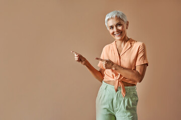 A modern stylish mature woman wearing an orange t-shirt and green pants of pastel shades is pointing with her fingers to the free space of the beige background on which she is standing. Copy space.