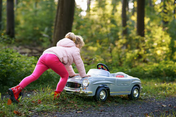 Naklejka na ściany i meble Little preschool girl driving big vintage toy car. Happy child having fun with playing outdoors. Active preschooler child enjoying warm autumn day in forest. Smiling stunning kid playing