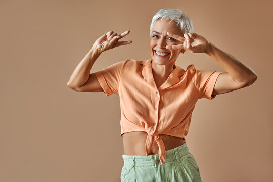 Beautiful positive modern mature confident smiling fun woman in orange t-shirt and green pants dancing on beige background. Excitement and pleasure.