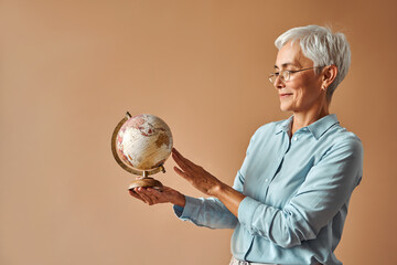Beautiful confident short haired gray haired woman mature teacher in blue blouse and glasses holds vintage globe and spins it. Studying, traveling around the world.