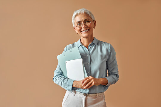 Modern confident beautiful gray-haired mature business woman in blue blouse and glasses holding laptop and work folders while standing on beige background and smiling.